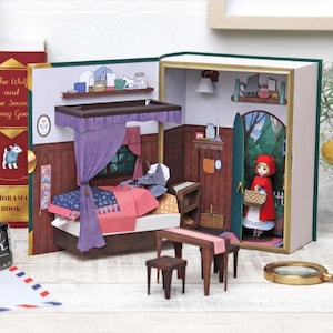 May include: A pop-up book diorama featuring a bedroom scene with a bed, table, and Little Red Riding Hood. The bed has a purple canopy and pink bedding. Red Riding Hood stands in a doorway holding a basket. The book is open.