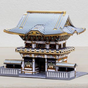 Può includere: Un modello di carta dettagliato di un tempio giapponese con un tetto a più livelli. Il modello presenta una combinazione di colori grigio e oro, con intricati dettagli architettonici. Il tempio è posto su una base grigia con una piccola recinzione.