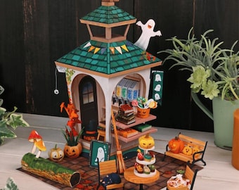 Op de afbeelding: Een miniatuur Halloween-diorama met een structuur met twee verdiepingen met een groen dak, een winkel, pompoenen, een spook, een bank, een tafel met lekkernijen en een bezem. De scène is in herfstkleuren.
