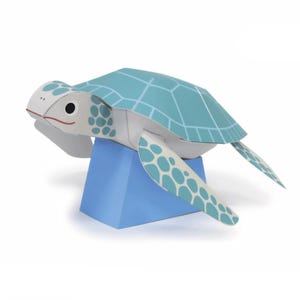 Può includere: Una tartaruga marina di carta nei toni del blu e del bianco, con un motivo geometrico sul guscio e segni a pois sulle pinne. La tartaruga è appoggiata su una base rettangolare blu, creando un effetto 3D.