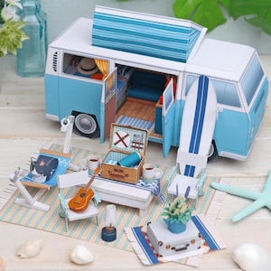 May include: A miniature blue and white camper van with an open door, revealing a detailed interior. A surfboard, picnic basket, and miniature furniture are arranged on a striped rug, creating a charming scene. A small guitar and a lamp are also present.
