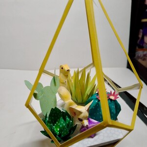 May include: A yellow geometric frame containing a paper craft diorama with a fennec fox and a prairie dog figurine. The scene includes various paper succulents in shades of green, a light green prickly pear cactus, and small pink and purple flowers.