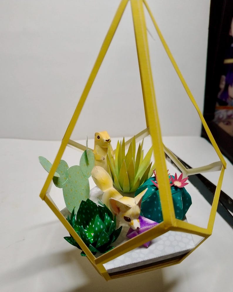 May include: A yellow geometric frame containing a paper craft diorama with a fennec fox and a prairie dog figurine. The scene includes various paper succulents in shades of green, a light green prickly pear cactus, and small pink and purple flowers.