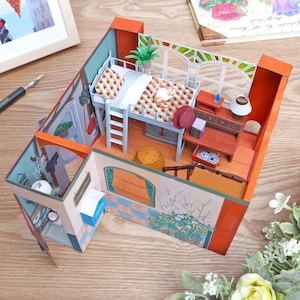 May include: A miniature paper dollhouse with a loft bed, desk, and window. The walls are painted in shades of orange, blue, and green. A small cat rests on the bed. The dollhouse is decorated with miniature furniture and accessories.