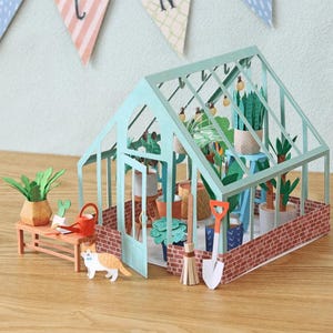 May include: A miniature paper greenhouse with a light green frame and brick-patterned base. Inside are various paper potted plants, a broom, and a shovel. Outside, a small wooden bench holds a potted plant, a watering can, and a paper cat figure.