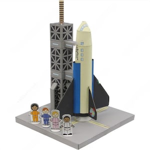 May include: A paper model of a space shuttle on a launchpad with four paper astronauts standing in front. The shuttle is white with blue and black accents. The launchpad is gray with a black base. The astronauts are wearing colorful spacesuits.