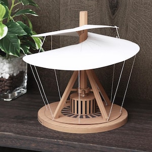 May include: A wooden model of a Leonardo da Vinci flying machine. The model features a circular base, a central column, and two white, curved wings. The design includes wooden supports and string detailing, showcasing a historical engineering concept.