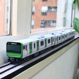 Puede incluir: Un tren en miniatura con una combinación de colores verde y blanco, colocado sobre una vía férrea en miniatura. El tren tiene varios vagones y está sobre una superficie blanca, con una ventana y un edificio al fondo.