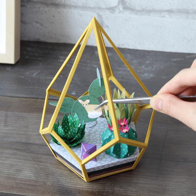 May include: A geometric gold-colored metal frame terrarium containing paper-crafted succulents and a small deer figure. The base features a gray patterned surface, and silver tweezers are positioned near the paper plants inside the structure.