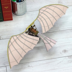 May include: A model of a Leonardo da Vinci flying machine. The wings are a light pink color with a series of parallel lines. The frame is made of wood and metal. The model is sitting on a light wood surface.