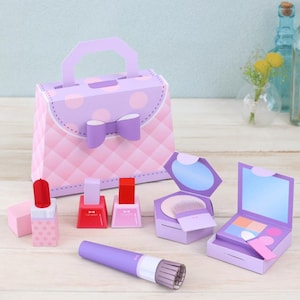 May include: A collection of paper makeup items, including a purse, lipstick, nail polish, a compact with a mirror and brush, and an eyeshadow palette. The items are primarily pink and purple, with a bow detail on the purse.