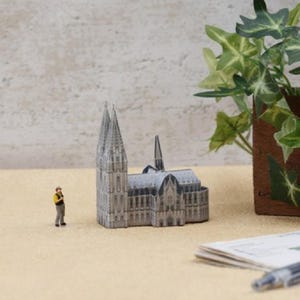 Könnte beinhalten: Ein graues Miniaturmodell einer Kathedrale mit zwei hohen Türmen und einem kleineren Turm. Eine kleine Figur in einem gelben Hemd und grauer Hose steht daneben. Das Modell steht auf einer beigen Oberfläche, mit Grün und einem Stift im Hintergrund.