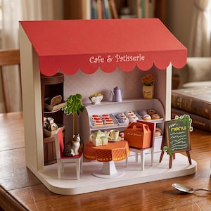 Może przedstawiać: Miniaturowa papierowa kawiarnia i cukiernia z czerwonym, falistym markizem z napisem Cafe & Patisserie. Wnętrze zawiera witrynę z małymi wypiekami, stolik z zestawem do herbaty, figurkę kota oraz tablicę z menu.