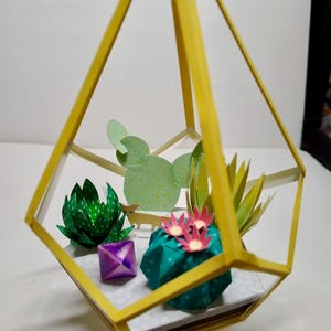 May include: A yellow geometric frame containing paper craft succulents and cacti. The arrangement includes a green spiky succulent, a teal cactus with pink flowers, a light green flat cactus, and a small purple cube. A tiny paper animal figure stands inside.