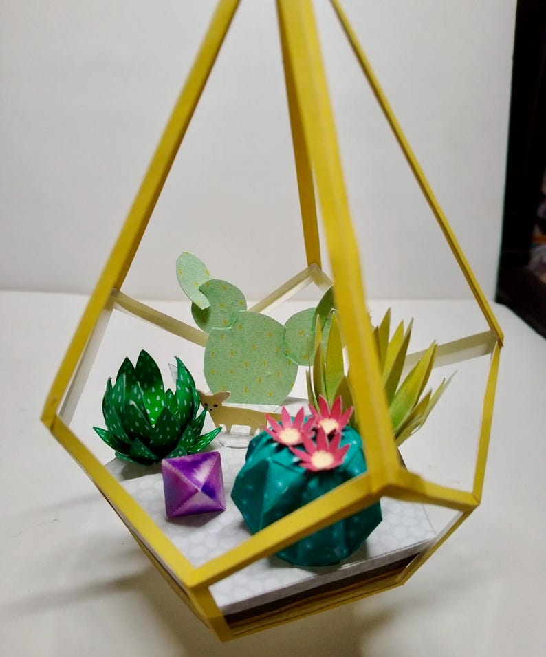 May include: A yellow geometric frame containing paper craft succulents and cacti. The arrangement includes a green spiky succulent, a teal cactus with pink flowers, a light green flat cactus, and a small purple cube. A tiny paper animal figure stands inside.