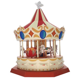 Könnte beinhalten: Ein Miniatur-Karussell mit einem rot-cremefarbenen Baldachin, verziert mit goldenen Akzenten und blauen und orangefarbenen Formen. Das Karussell verfügt über mehrere weiße Pferde und wird von einer kleinen Engelsfigur gekrönt. Der Sockel ist rot.