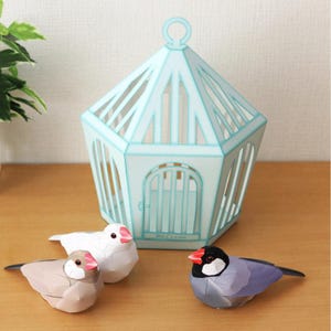 Pode incluir: Uma gaiola de pássaros de papel azul claro com uma alça redonda e uma porta pequena, acompanhada por três pássaros de papel em branco, cinza e azul. Os pássaros têm bicos vermelhos e marcas pretas. A palavra "Welcome" está impressa na gaiola.