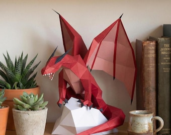 Puede incluir: Una escultura de dragón de papel rojo con detalles en negro, posada sobre una base geométrica blanca. El dragón tiene grandes alas y una larga cola. La escultura se exhibe en una estantería de madera con plantas en macetas y libros.
