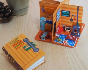 May include: A miniature pop-up book titled "LOG CABIN" with a detailed interior scene. The open book reveals a cosy cabin interior with a fireplace, furniture, and a blue rug. The cover is brown and yellow with a silver handle.