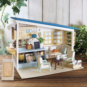 May include: A miniature paper model of a cafe with a blue roof, featuring a counter with food, a menu board, and seating areas. The cafe includes a coffee sign, tables, chairs, and decorative elements.