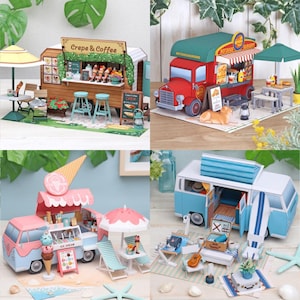 Puede incluir: Una colección de cuatro dioramas en miniatura de camiones de comida y caravanas. El conjunto incluye un puesto de crepes y café de madera, un camión rojo de perritos calientes y hamburguesas, una furgoneta de helados rosa y una caravana azul con accesorios.