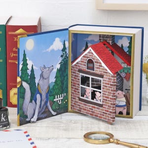 Könnte beinhalten: Ein Pop-up-Buch, das die Geschichte der drei kleinen Schweinchen darstellt. Das offene Buch zeigt einen Wolf, ein Backsteinhaus und drei Schweine. Das Haus hat ein rotes Dach und einen Schornstein. Der Wolf blickt in den Himmel.