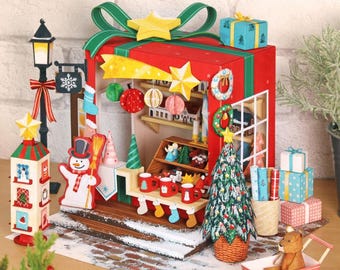 May include: A miniature Christmas village scene with a red gift box-shaped building, a snowman, and a Christmas tree. The scene includes a street lamp, presents, and a small sled with a toy animal. The color palette is red, green, and white.