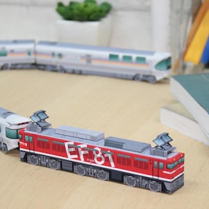 May include: A red and gray model train with the text "EF 81" on the side. The model train is on a wooden surface, with other train cars in the background. The model train has detailed features.