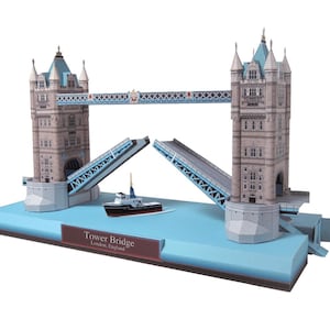 Könnte beinhalten: Ein Papiermodellbausatz der Tower Bridge in London, England. Die Brücke ist aus weißem Papier mit blauen Akzenten und wird mit der Zugbrücke hochgefahren gezeigt. Das Modell steht auf einem blauen Sockel mit der Aufschrift "Tower Bridge, London, England".
