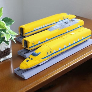 Può includere: Tre modellini di treni gialli con strisce blu sono esposti su binari grigi. I treni sono disposti su una superficie di legno, con una pianta in un vaso decorativo visibile sullo sfondo.