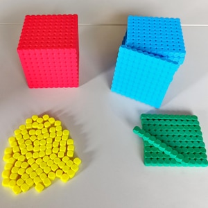 Puede incluir: Cuatro manipulativos matemáticos coloridos: un cubo rojo, un cubo azul, un montón de piezas amarillas y un cuadrado verde con un palo. Los cubos están hechos de pequeños bloques conectados. Las piezas amarillas son pequeñas y redondeadas.