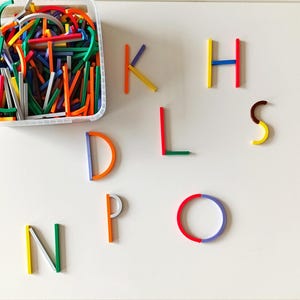 May include: A clear plastic container filled with colorful wooden sticks and curved pieces. Several letters of the alphabet are arranged on a white surface, constructed from the same colorful wooden pieces. The letters include D, H, K, L, N, O, P, and S.
