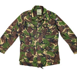 Pode incluir: Casaco de camuflagem em tons de verde, castanho e bege. O casaco apresenta vários bolsos, fechos de botão e um pequeno emblema da bandeira britânica na manga. Gola e mangas compridas.