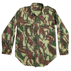 Puede incluir: Camisa de camuflaje de manga larga en tonos verdes, marrones y beige. La camisa tiene dos bolsillos en el pecho, cierre de botones y charreteras en los hombros. La tela presenta un patrón de pinceladas.