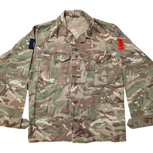 Könnte beinhalten: Hemd mit Tarnmuster, Knopfleiste, zwei Brusttaschen und geknöpften Manschetten. Das Hemd hat Aufnäher auf den Ärmeln und einen Kragen. Die Farbpalette umfasst Grün-, Braun- und Beigetöne.