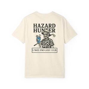 May include: Cream-colored t-shirt featuring a graphic of a skeleton golfer with a drink and golf club. The text "HAZARD HUNTER" is above the image, and "LAKELAND GOLF CLUB" is below. The skeleton wears a patterned shirt and a golf hat.