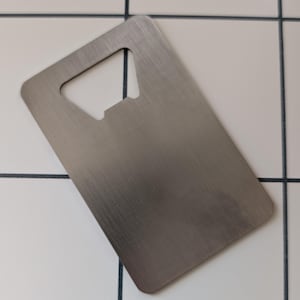 May include: A brushed metal bottle opener in a rectangular shape with a bottle cap opener at the top. The opener is silver and has a smooth, clean surface. The background is a white tile.
