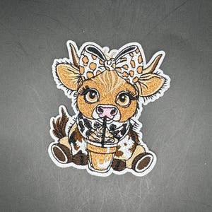 Op de afbeelding: Geborduurde patch met een schattige cartoonkoe met een strik, sjaal en een drankje. De koe is bruin, wit en zwart met grote ogen. De strik heeft een luipaardprint. Het drankje zit in een doorzichtige beker met een rietje.