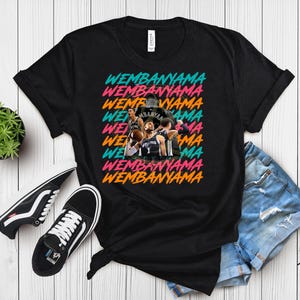 May include: Black t-shirt with the word "WEMBANYAMA" in a repeating, colorful design. The shirt features a team photo in the center. A pair of black and white sneakers and a small potted plant are also visible.