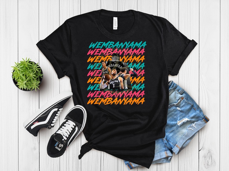 May include: Black t-shirt with the word "WEMBANYAMA" in a repeating, colorful design. The shirt features a team photo in the center. A pair of black and white sneakers and a small potted plant are also visible.