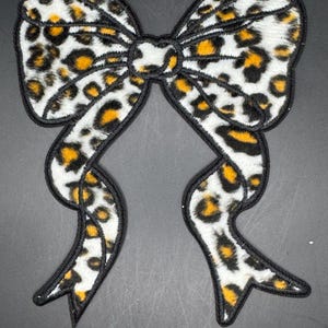 May include: A decorative bow patch with a leopard print pattern. The bow features a white background with black and yellow spots, outlined with black stitching. The bow's design includes two loops and two flowing tails.