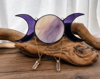 May include: A stained glass triple moon symbol featuring a central circular piece with white and purple swirls, flanked by two dark purple crescent moons. Two gold-toned metal chains hang from the bottom, each holding a clear, pointed crystal pendant.