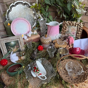 May include: A collection of vintage items, including a silver tray, pink plates, a floral wreath, a pink pitcher, and a teacup with a strawberry design. Decorative elements include small glass vases, a gold picture frame, and a woven basket.
