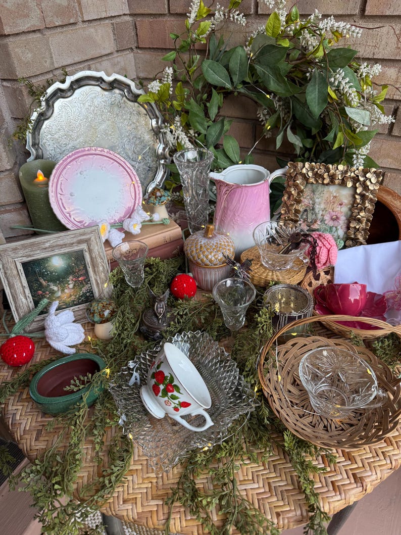 May include: A collection of vintage items, including a silver tray, pink plates, a floral wreath, a pink pitcher, and a teacup with a strawberry design. Decorative elements include small glass vases, a gold picture frame, and a woven basket.