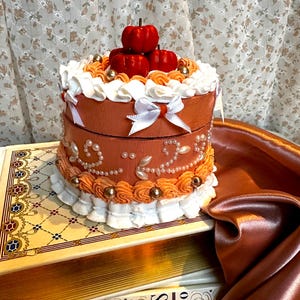 May include: A two-tiered, round, orange and white decorative box with a white ribbon and three red pumpkins on top. The box is sitting on top of two books.