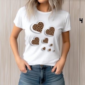 May include: White t-shirt featuring a design of several heart shapes with a giraffe print pattern. The hearts vary in size and are arranged to appear as if they are floating. The shirt is a crew neck style.