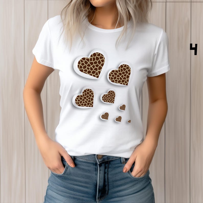 May include: White t-shirt featuring a design of several heart shapes with a giraffe print pattern. The hearts vary in size and are arranged to appear as if they are floating. The shirt is a crew neck style.