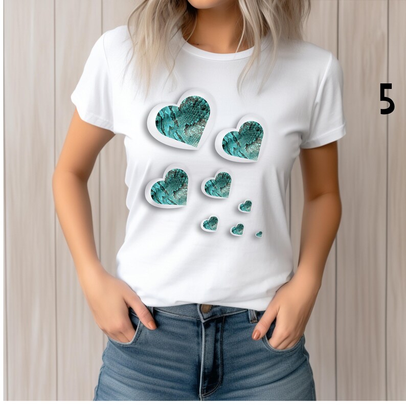 May include: White t-shirt featuring a design of several hearts in varying sizes. The hearts have a teal snakeskin pattern and a white border. The shirt is paired with blue denim jeans.