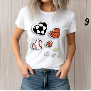 May include: White t-shirt featuring heart-shaped sports ball graphics: a soccer ball, basketball, baseball, and football. Additional small heart graphics include a volleyball and a tennis ball. The number 9 is visible on the right side.