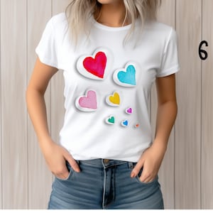 May include: White t-shirt featuring a watercolor design of colorful hearts in red, blue, pink, yellow, and green. The hearts have a white outline and a 3D effect. The shirt is worn by a person in blue jeans.